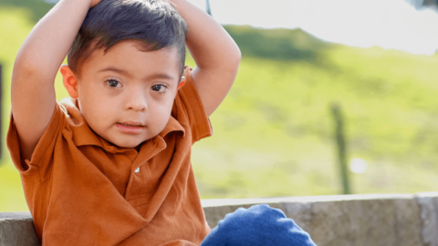 Diagnoses (1) Young boy wearing an orange shirt sits outdoors on a stone bench with his arms raised above his head, looking slightly to the side.