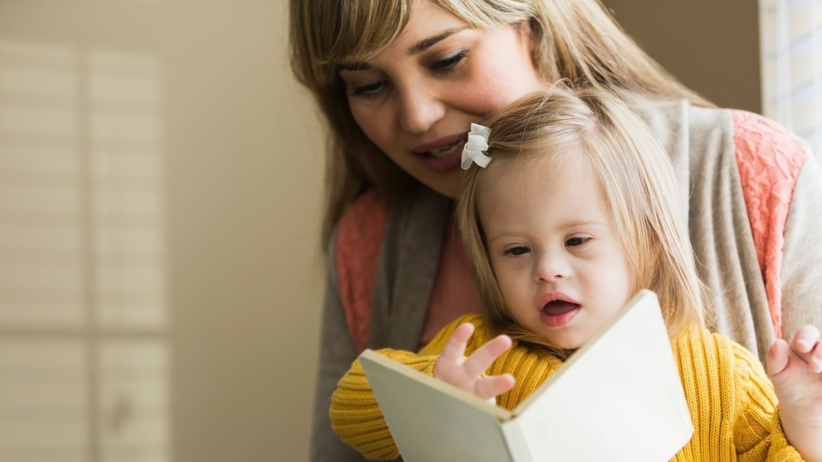 canchild-banner An adult and a young child with Down syndrome read a book together indoors; the child is wearing a yellow sweater and a white bow.