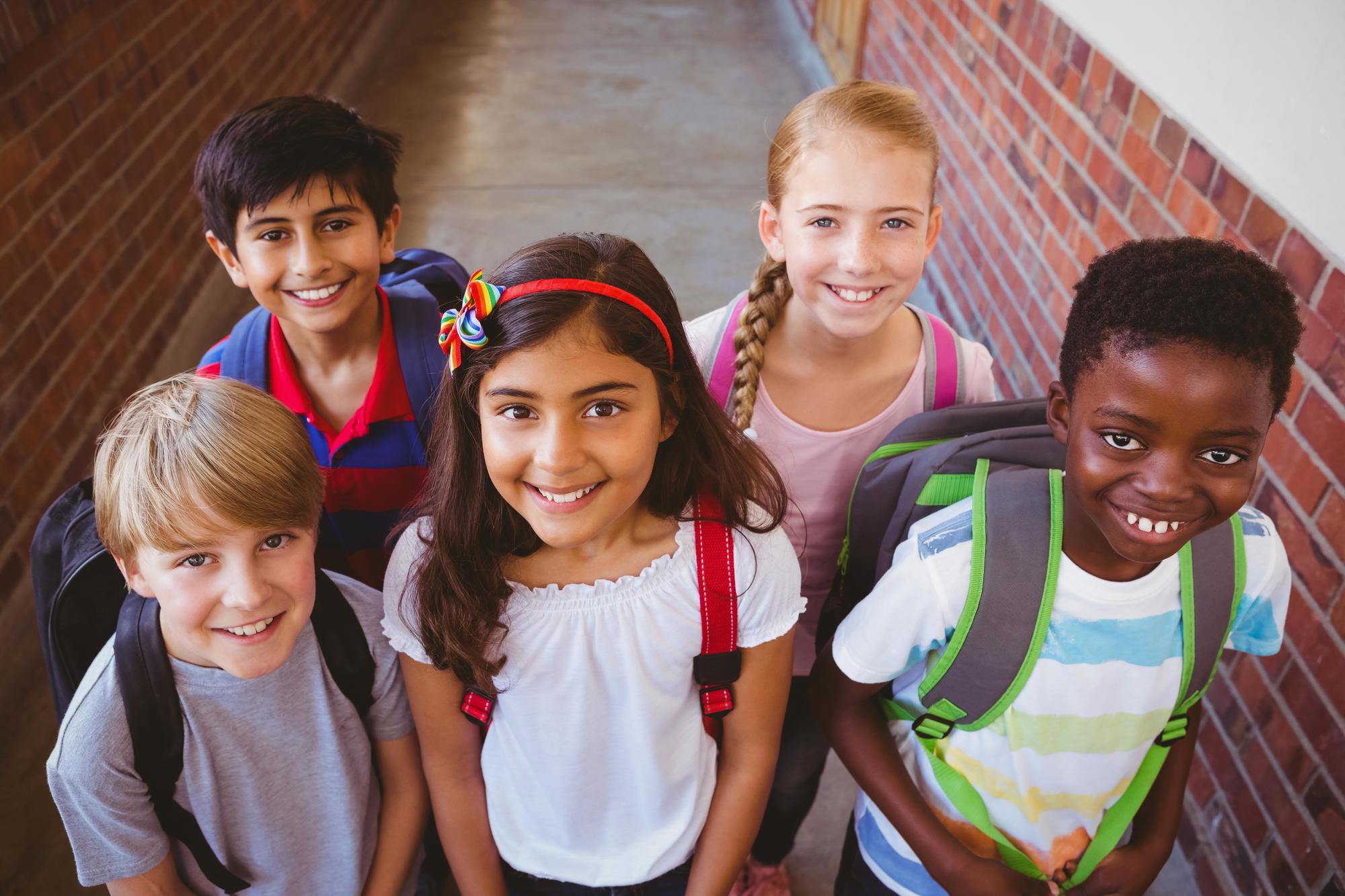 5_children_smiling_at_camera Five smiling children with backpacks stand together in a school hallway, facing the camera.