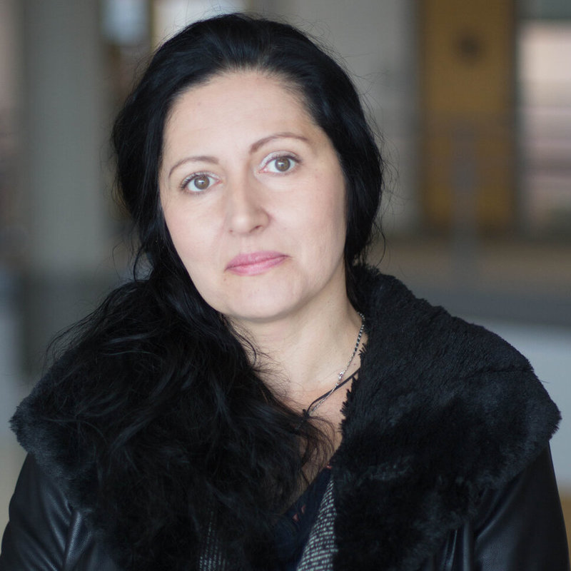 Danijela_Grahovac A woman with long dark hair and a black coat looks at the camera with a neutral expression in an indoor setting.