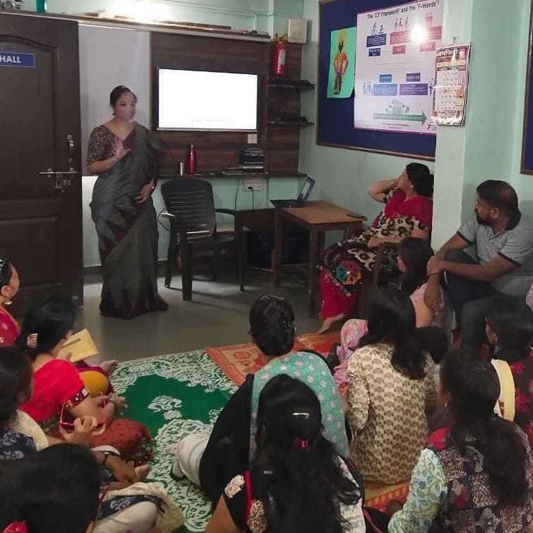 F-Words-India3 A woman stands near a screen giving a presentation to a seated group of adults and children in a small room. Educational posters are visible on the walls.