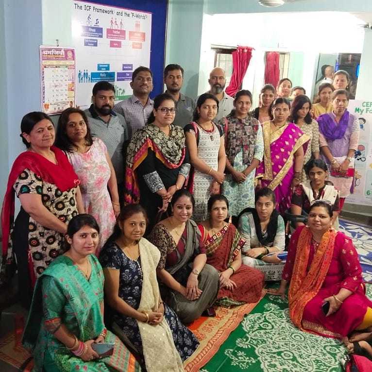 FWords-India A group of men and women pose for a photo indoors, with posters and charts on the walls, some seated on the floor and others standing behind.