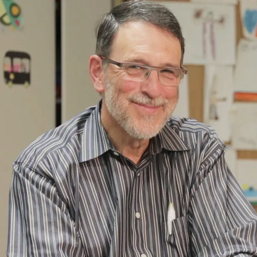 Peter A man with gray hair, glasses, and a beard smiles while wearing a striped shirt; drawings are visible on a wall in the background.
