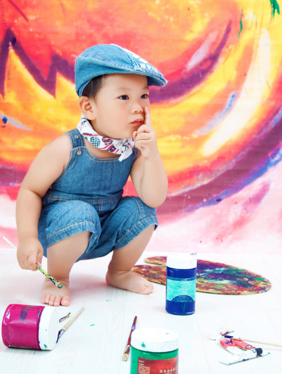 child-1 A young child in denim overalls and cap squats in front of a colorful painted backdrop, holding a paintbrush with art supplies scattered on the floor.