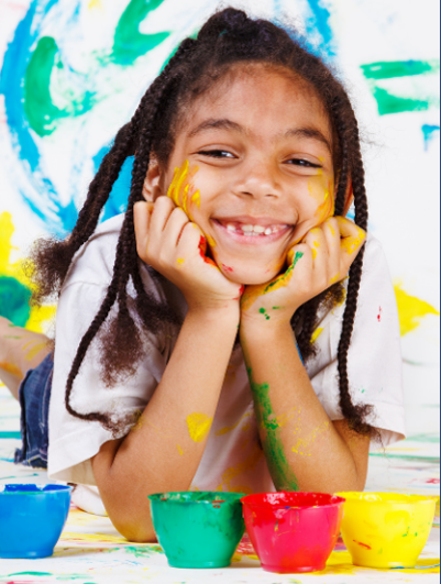 child-3 Smiling child with painted hands and face lies in front of colorful paint cups, with a painted background behind her.