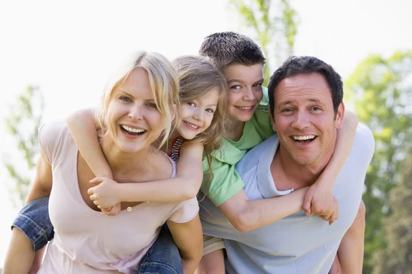 depositphotos_4767995-stock-photo-couple-giving-two-young-children Two adults and two children are outdoors, smiling. The children are on the adults’ backs, and they are all looking at the camera. Trees and sky are visible in the background.