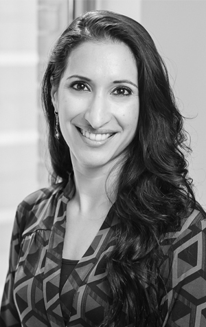 Amaria-cropped A woman with long dark hair smiles at the camera, wearing a patterned top and earrings, standing indoors in front of a window.