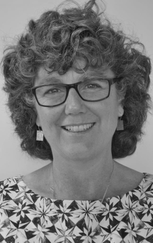 Christine-Imms-2020 Black and white portrait of a smiling woman with curly hair, wearing glasses, geometric earrings, and a patterned top.