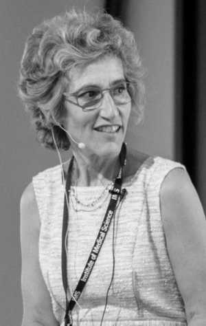 A woman with curly hair and glasses, wearing a sleeveless dress and a lanyard, speaks into a headset microphone.