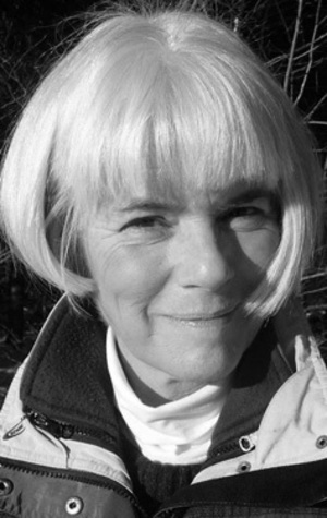 A woman with short, light-colored hair and a bob haircut, wearing a jacket and turtleneck, smiles slightly while outdoors. The photo is in black and white.