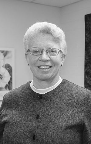 Doreen_Bartlett_(2) A person with short, light-colored hair and glasses, wearing a buttoned sweater, stands indoors and smiles at the camera.