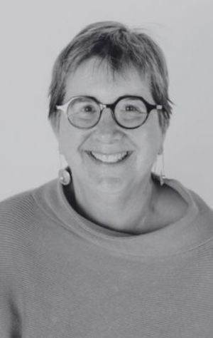Ginny_Paleg A person with short hair and glasses smiles at the camera, wearing a light-colored top and earrings, against a plain background.