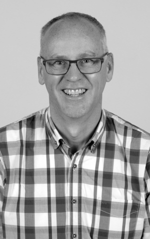 A middle-aged man with short hair and glasses smiles at the camera. He is wearing a plaid button-up shirt and is photographed against a plain background.