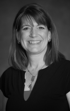 HURTUBISE_karen_2022 Black and white portrait of a woman with shoulder-length hair, smiling, wearing a dark top and a necklace, with a neutral background.