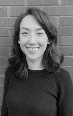 Hanae A woman with shoulder-length dark hair and a black sweater smiles in front of a brick wall. The image is in black and white.