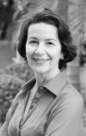 Livia Black and white photo of a woman with short dark hair, wearing a button-up shirt, smiling, and standing outdoors with trees in the background.