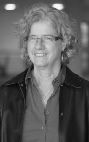 Marilyn-Wright A woman with curly hair and glasses is smiling. She is wearing a collared shirt, a jacket, and a necklace. The image is in black and white.