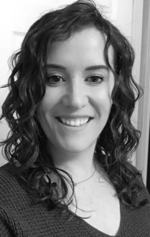 Meaghan_Reitzel A woman with wavy hair smiles at the camera in a black and white photo, standing indoors in front of a door.