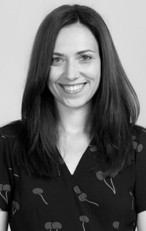 Monika-Novak A woman with straight, dark hair and a patterned dark top smiles at the camera against a plain background.