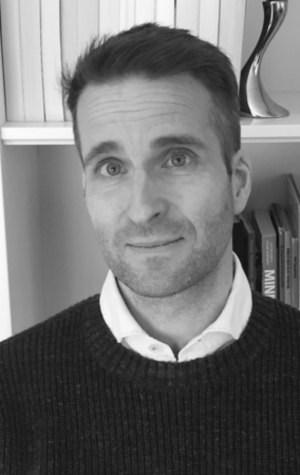 Olaf-V Black and white photo of a man with short hair and a trimmed beard, wearing a collared shirt and sweater, standing in front of shelves with books and decorative items.