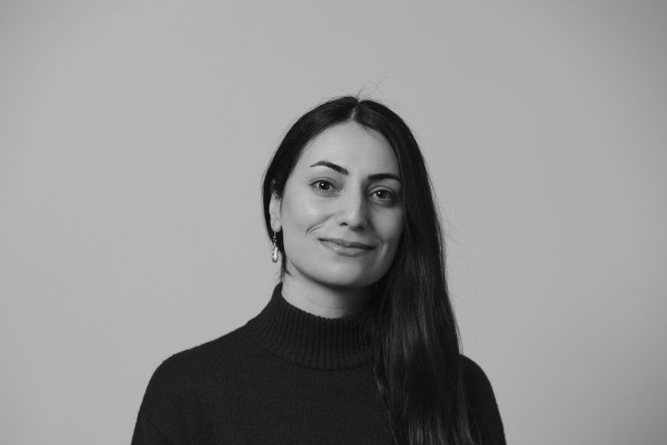 A woman with long dark hair, wearing a turtleneck sweater and earrings, smiles slightly while looking at the camera against a plain background. Black and white photo.