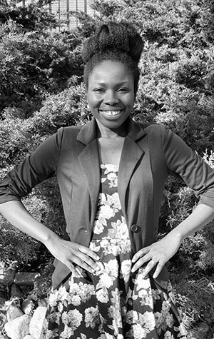 Photo_Christiana A woman stands outdoors in front of bushes, smiling at the camera. She wears a floral dress and a blazer, with her hands on her hips. The image is in black and white.