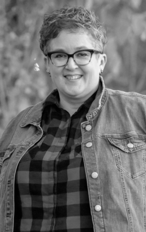 A person with short curly hair, glasses, and a denim jacket over a plaid shirt, smiling outdoors.