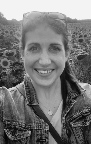 A woman wearing a denim jacket and glasses on her head smiles at the camera in front of a sunflower field.
