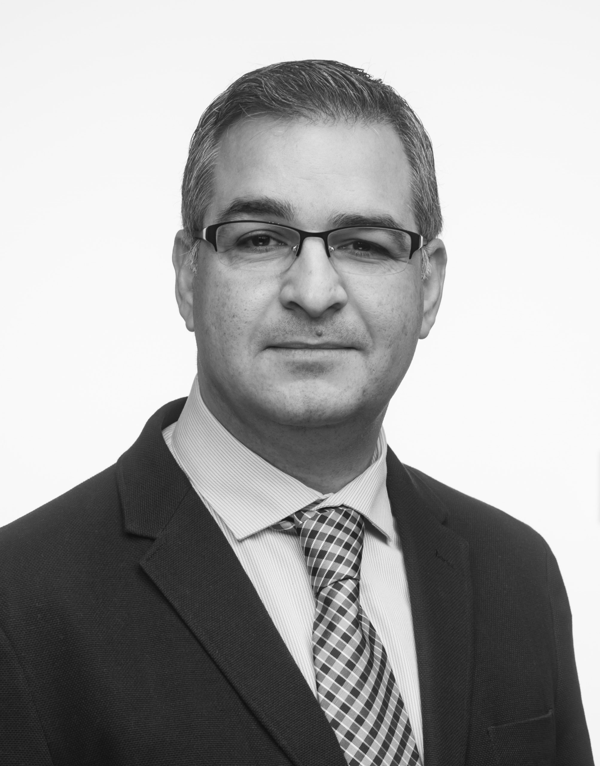 A man wearing glasses, a suit, and a checkered tie poses for a professional headshot against a plain background.