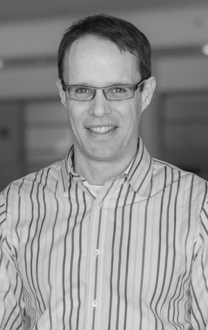 StephenGentles A man wearing glasses and a striped shirt smiles at the camera in an indoor setting. The photo is in black and white.