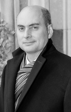 Steven_Hanna_cropped A man with a bald head and wearing a dark coat and striped scarf stands in front of a stone wall. The image is in black and white.