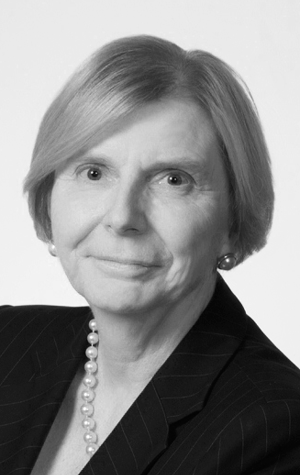 Portrait of an older woman with short light hair, wearing a dark suit, pearl necklace, and pearl earrings, facing the camera with a neutral expression.