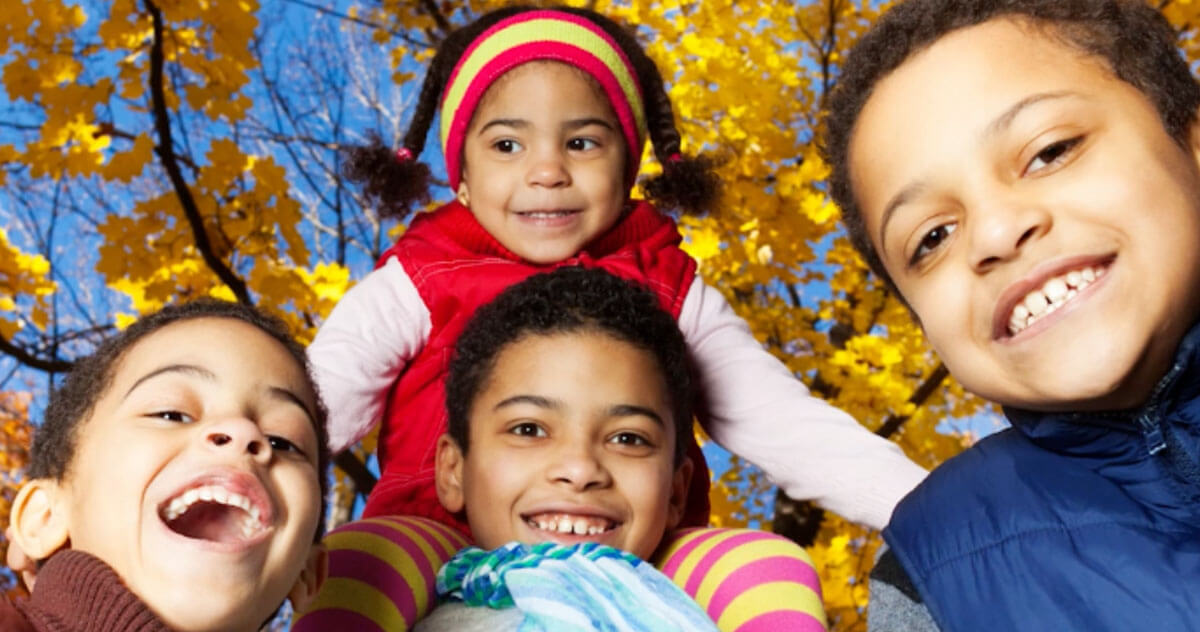 Untitled-1 Four children smile outdoors in front of bright yellow autumn leaves, wearing warm clothing such as jackets, hats, and scarves.