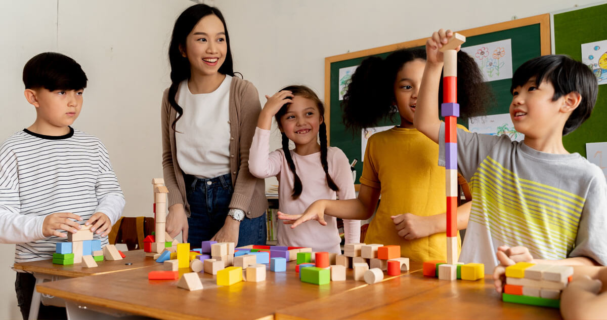 Untitled-2 Four children and a teacher interact with wooden building blocks at a classroom table; one child is stacking blocks into a tall tower.