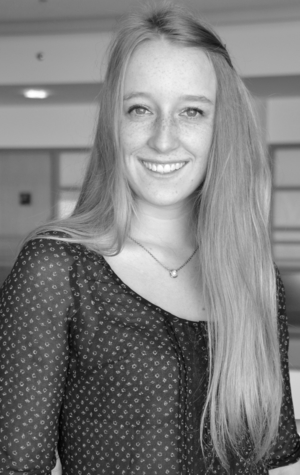 A woman with long straight hair, wearing a patterned blouse and necklace, smiles at the camera in an indoor setting. The image is in black and white.
