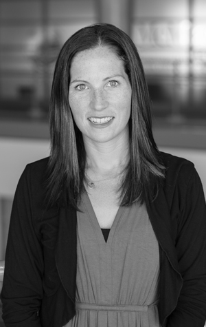 A woman with straight, shoulder-length hair wearing a dark jacket over a dress, standing indoors with a blurred background.