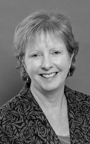missiuna_croppedBW2 Black and white portrait of a middle-aged woman with short hair, wearing a patterned jacket, smiling in front of a plain background.