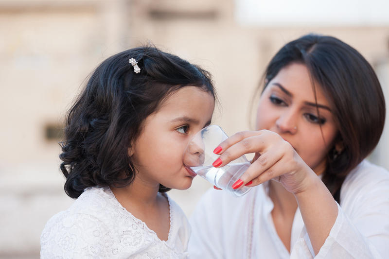Bringing_More_Diverse_Family_Voices_feature A woman helps a young girl drink water from a glass, both dressed in white, with a neutral background.
