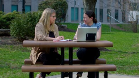Get Involved (1) Two women sit across from each other at an outdoor picnic table, engaged in conversation; one has a laptop and the other has an open notebook.