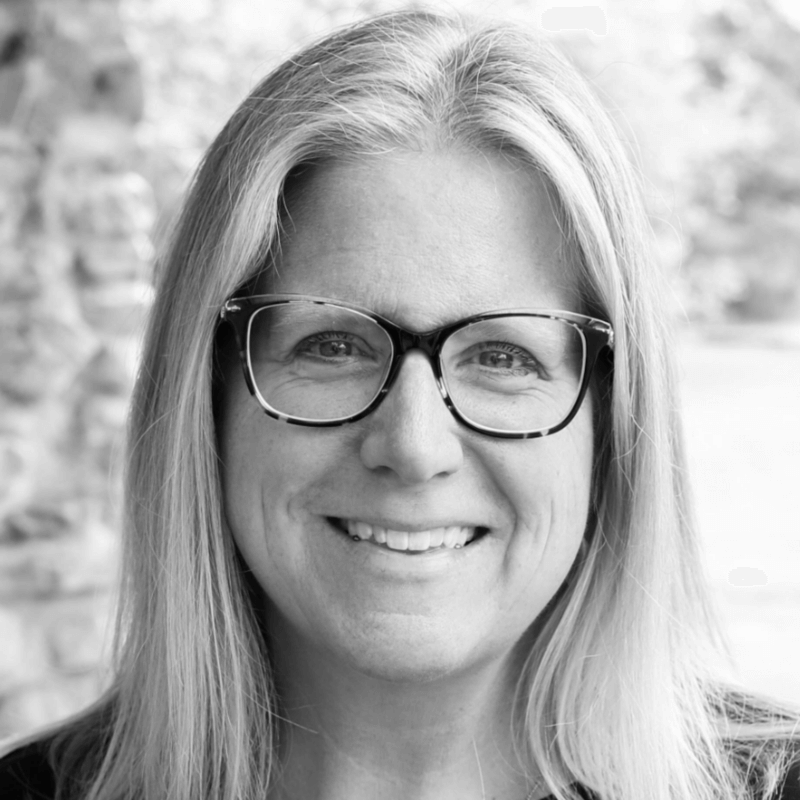 A person with long light hair and glasses is smiling, facing the camera. The image is in black and white with a blurred outdoor background.