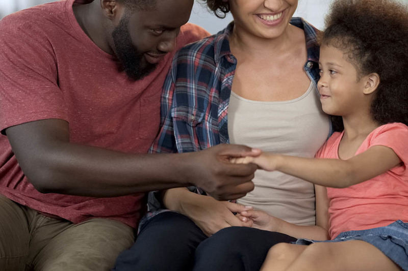 The_Importance_of_Family_Engagement_in_Research A man, woman, and young girl sit closely together on a couch, smiling and holding hands.