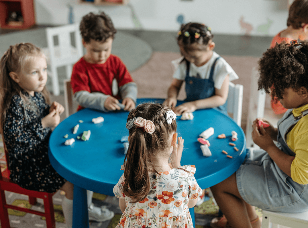 Five young children sit around a blue table, playing with modeling clay in a classroom setting.