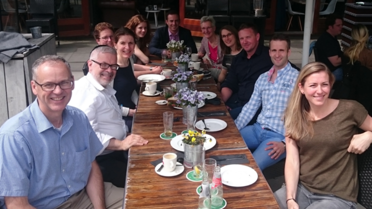 A group of people sit around a long outdoor table with drinks and flowers, smiling at the camera in a sunny café setting.