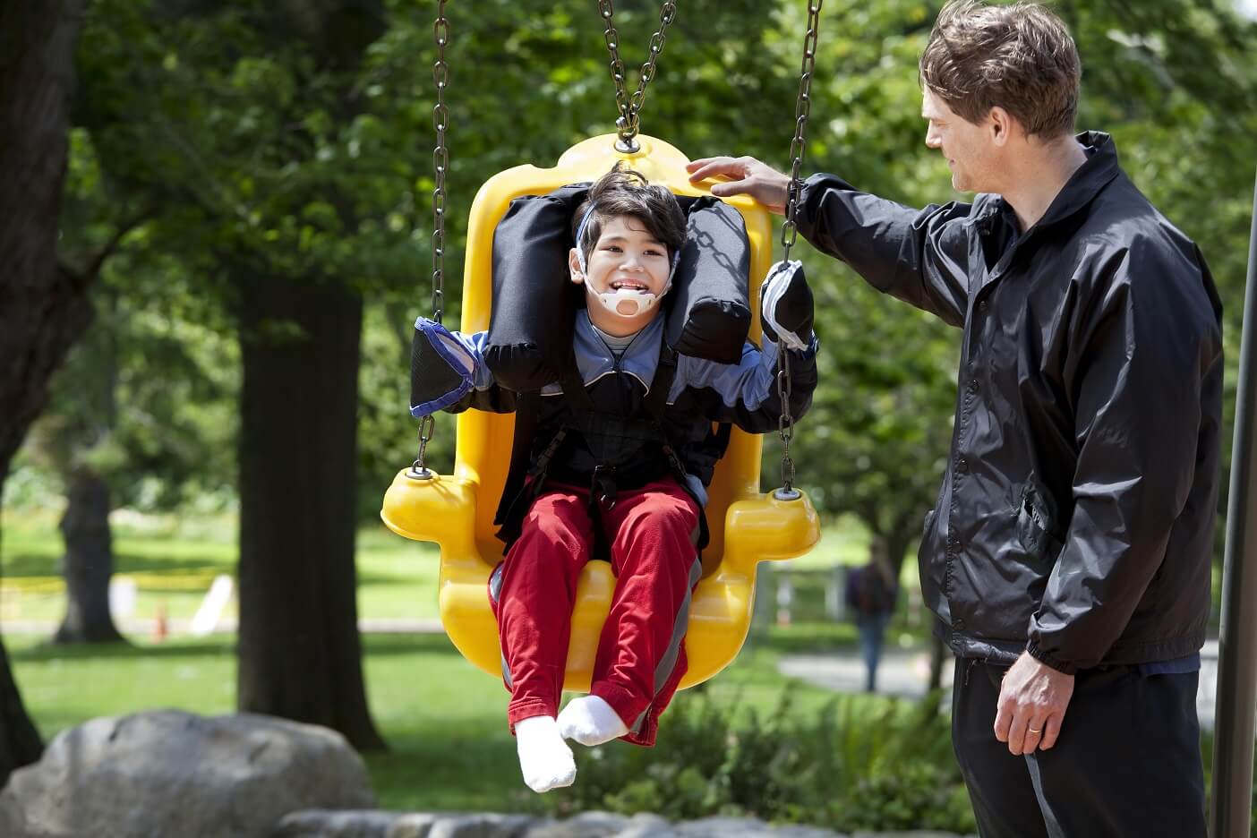 A child with medical support equipment sits on an adaptive swing, smiling, while an adult stands nearby in an outdoor park setting.