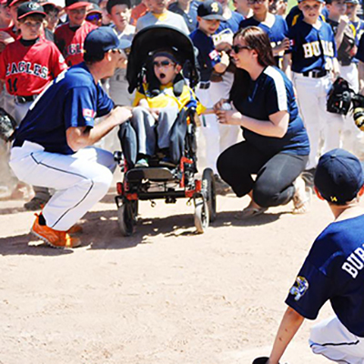 Fml116w A baseball coach and woman interact with a child in a wheelchair on a baseball field, surrounded by young players and spectators.