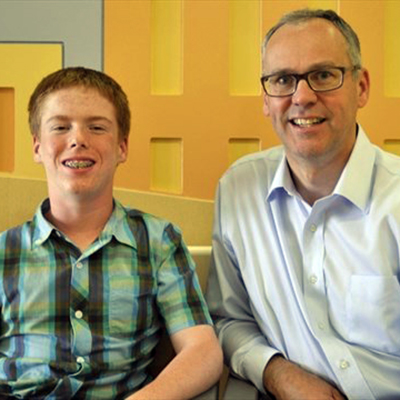 JbY3WAu Two men, one younger with braces and wearing a plaid shirt, and one older in glasses and a light dress shirt, sit side by side in front of a yellow wall.