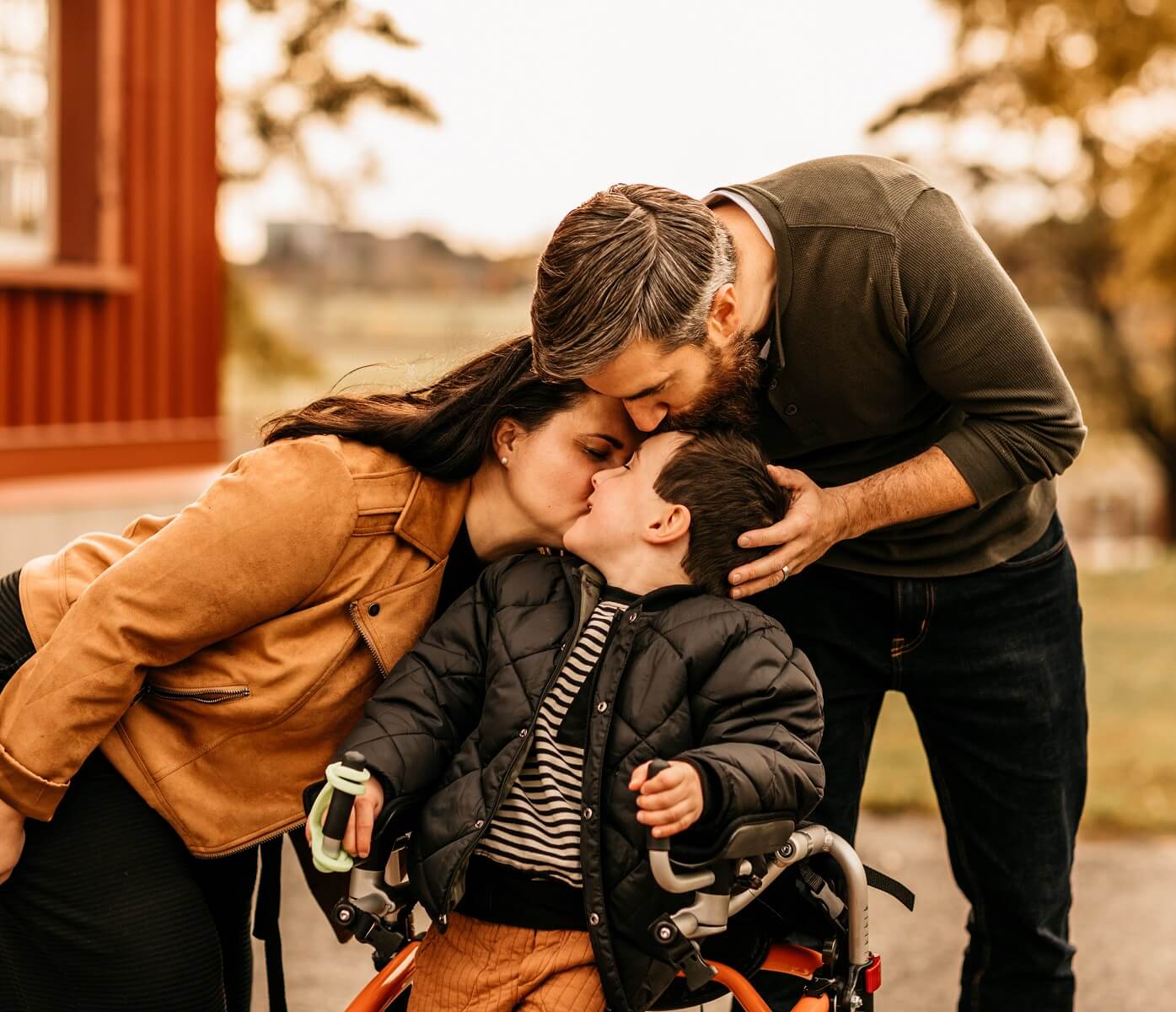 Of_Wild_Dawn_Photography-_Ottawa_Ontario__Crystal_Dawn_Cook-0670 (1) A child in a wheelchair is kissed on the forehead and cheek by two adults outdoors, with autumn trees in the background.