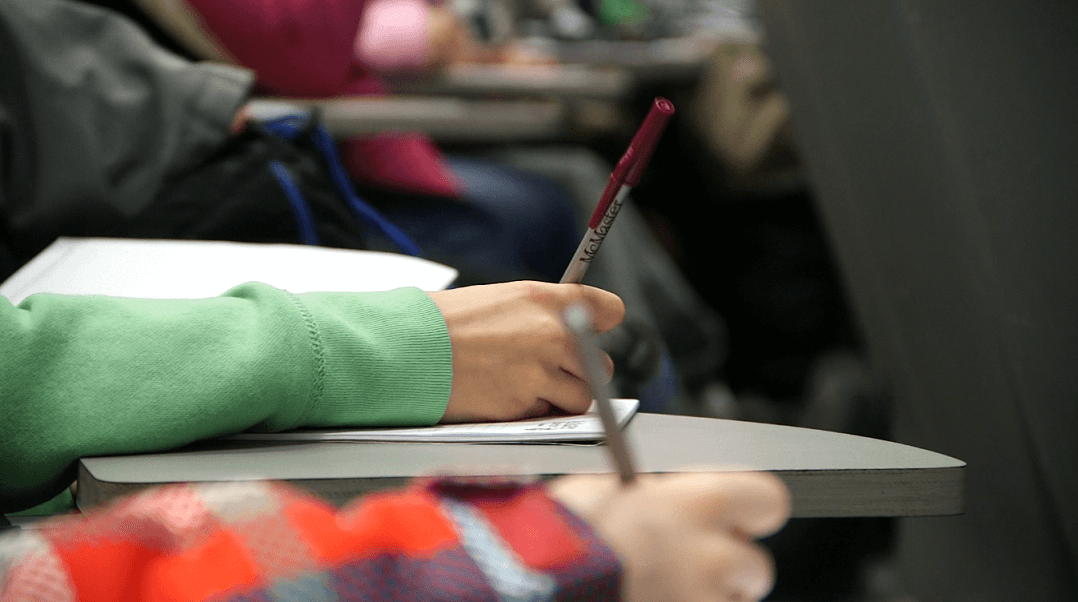 Close-up of two people writing in notebooks at desks, one in a green sleeve and the other in a plaid sleeve, in what appears to be a classroom or lecture hall setting.