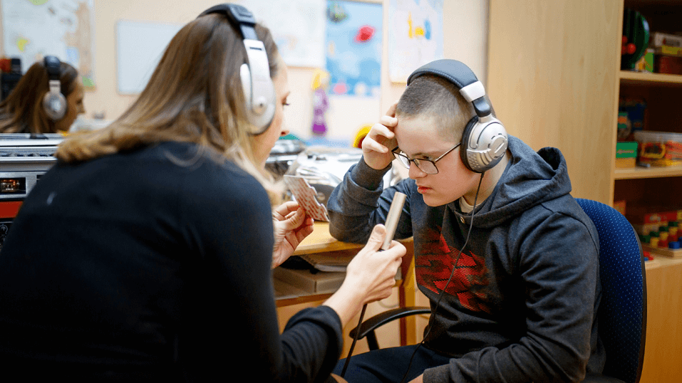 Untitled design-25 (1) A young person wearing headphones sits and listens as an adult holds a microphone or recorder toward them in a classroom or therapy setting.
