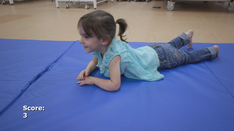 A young girl in a blue top and jeans lies on her stomach on a blue mat with the text "Score: 3." Featured in the Gross Motor Function Measure Training Video.